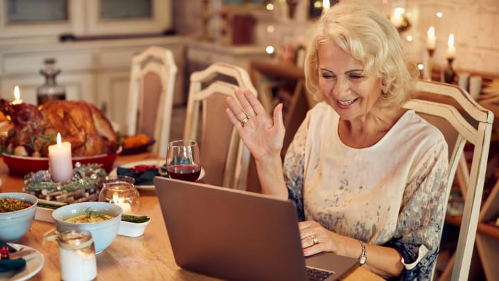 Elderly woman waving at an open laptop surrounded by a glass of wine, a large roasted turkey, and other food.