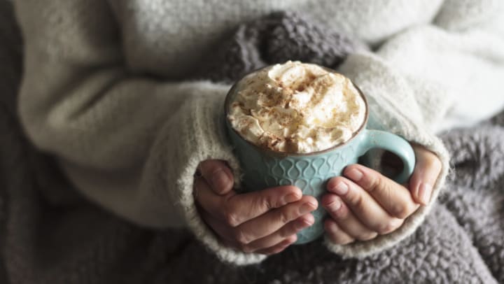 Person in a sweater with a blanket on their lap holding a light blue mug of hot chocolate with whipped cream.
