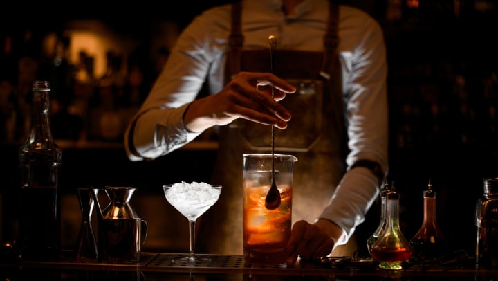 A bartender in white shirt and leather apron stirring an alcoholic cocktail in a glass shaker, with a special bar spoon.