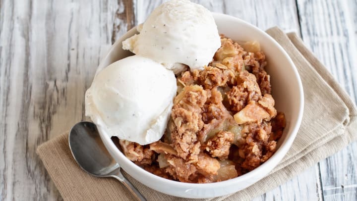 A bowl of apple crumble with two scoops of ice cream sitting on a folded napkin with a spoon next to it. 