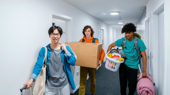 Three young male students carrying boxes and belongings through a dorm hallway.