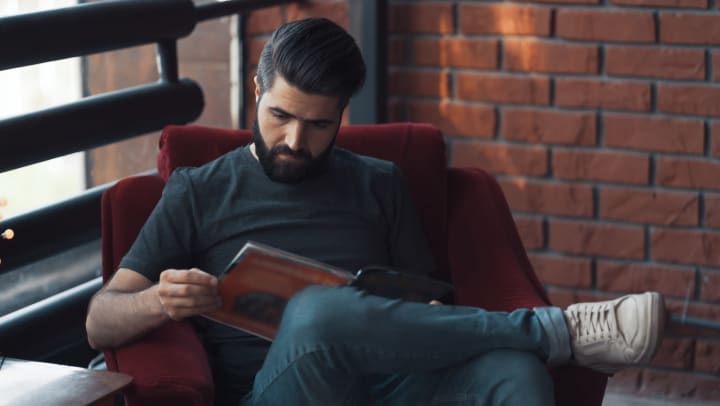 A bearded man sitting in a chair reading a graphic novel or magazine.