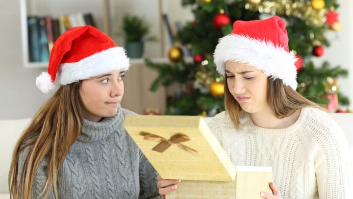 Two women with Santa hats sitting on a couch. One woman is holding an open gift box and making a distressed face.