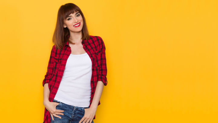 Woman wearing a red flannel button down with a white shirt underneath while posing in front of a yellow wall. 