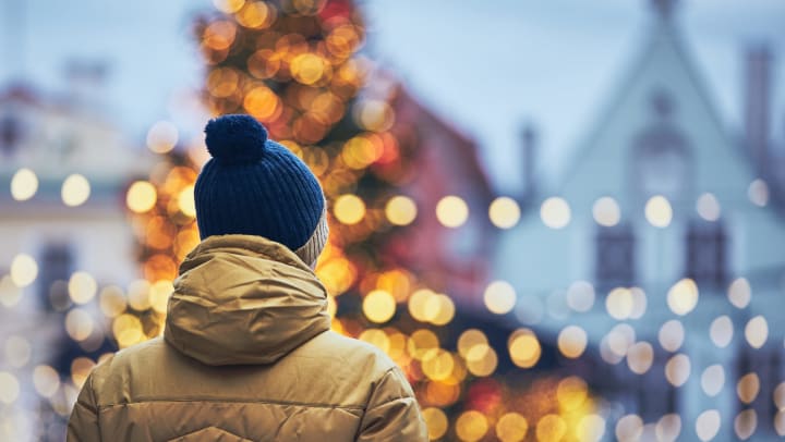The back of a person in a yellow coat and skull cap with a big Christmas tree in the background. 