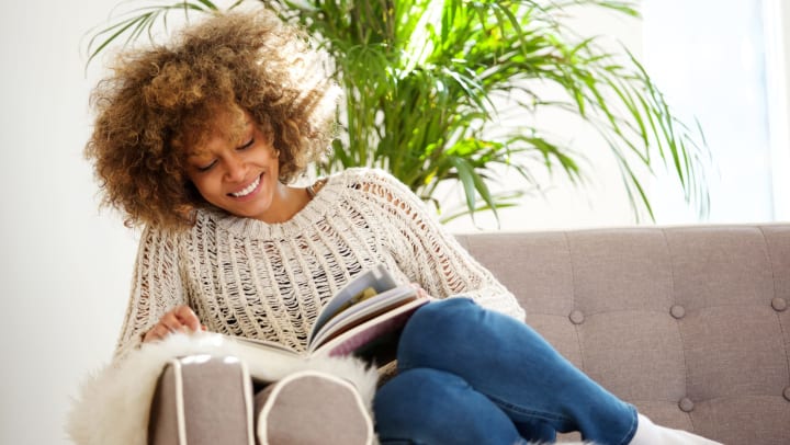 A smiling woman sits on a sofa looking at a hardcover book.