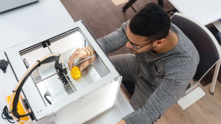 Aerial view of a man with his hand in a 3D printer, touching a yellow object.