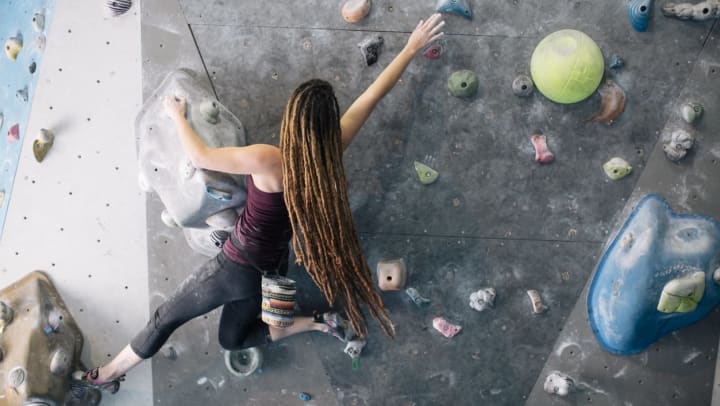 A person with long hair reaches between hand holds while climbing a wall at an indoor climbing gym