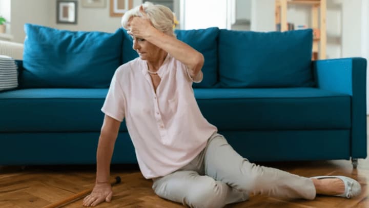 An older woman sitting on the floor with her hand on her forehead, looking left.