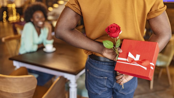 A man hides a red rose and a wrapped Valentine’s Day gift behind his back as he approaches his smiling girlfriend who is seated at a table in a restaurant. 