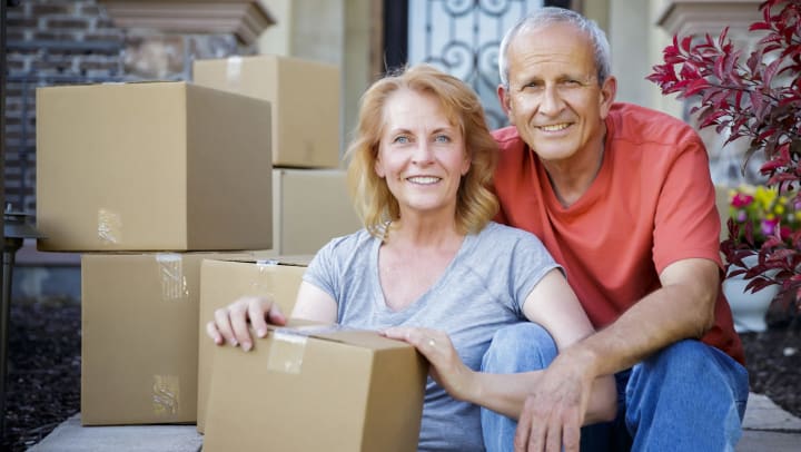 A mature couple sitting on the steps in front of their home with moving boxes.