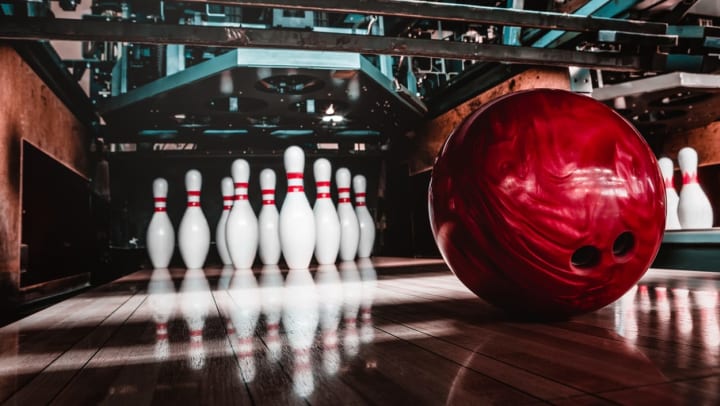 An action shot of a deep red, marbled bowling ball rolling down the lane toward a fresh frame of pins