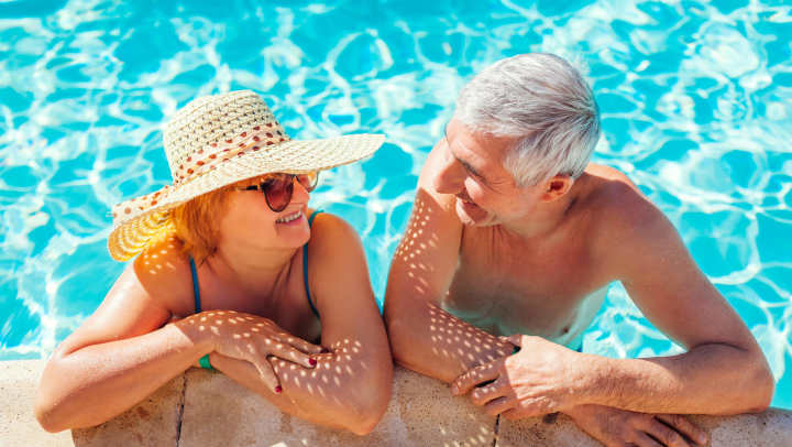 A senior couple smiling together by a sparkling outdoor pool on a sunny day.