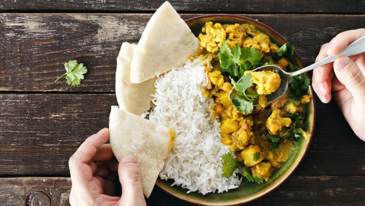 Overhead view of a man’s hands holding a spoon and a piece of naan bread above a plate of spicy cauliflower curry with rice. 