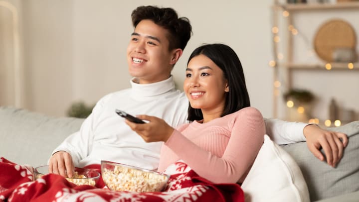 A man and a woman sitting on a couch eating popcorn. The woman is pointing a TV remote. 