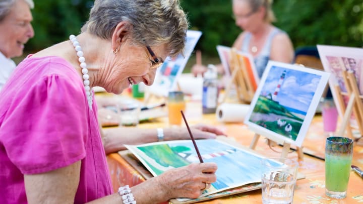 An elderly woman smiling while painting a picture outdoors surrounded by other women painting.