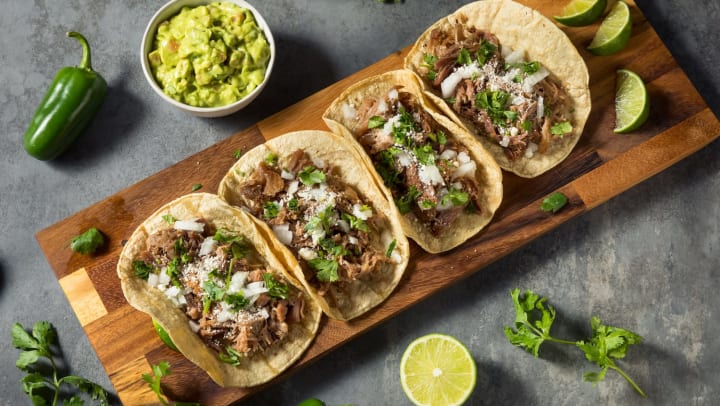 An overhead shot of a wood tray topped with four carnitas tacos with onion and cilantro, served with lime wedges and a bowl of guacamole
