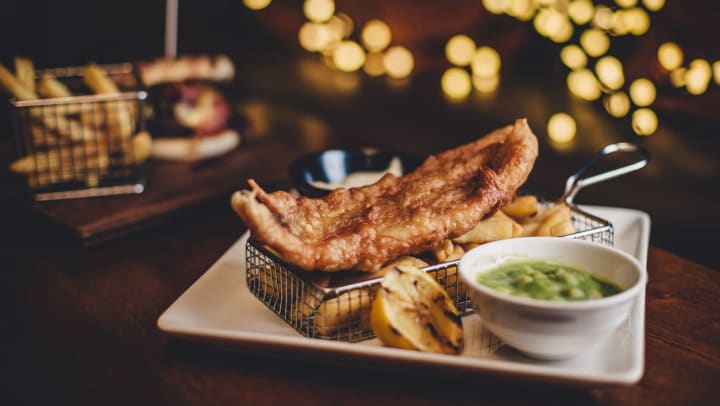 A small metal frying basket containing fresh fries topped with a piece of breaded fish, served with a lemon wedge and a side cup of mushy peas