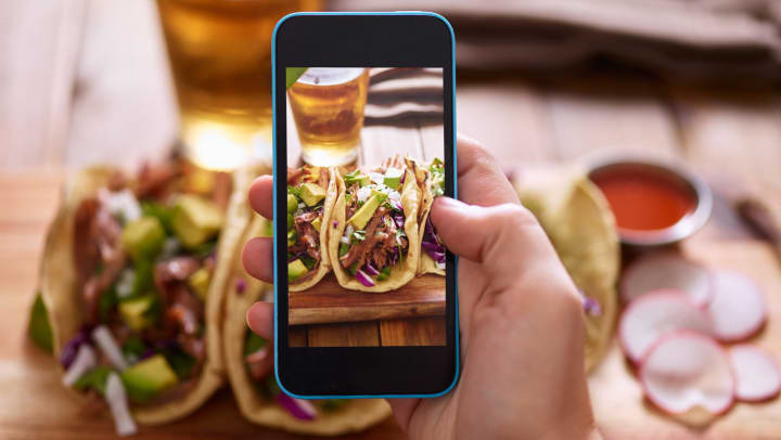 View of a hand holding a smartphone and taking a picture of a plate of tacos and a glass of beer