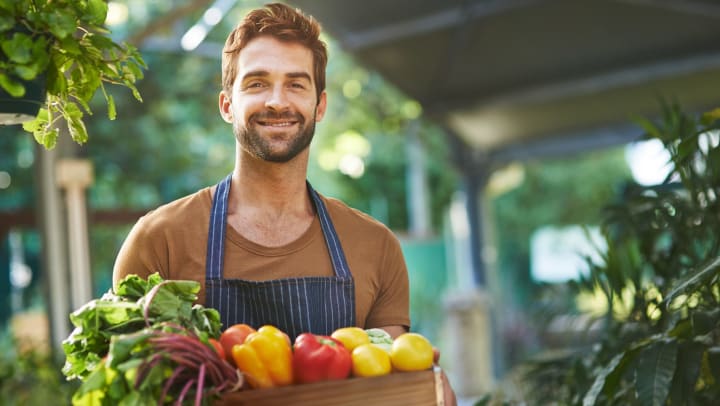 Man wearing an apron and holding a box of fresh vegetables, smiling toward the camera.