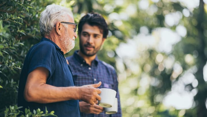 Adult son having a conversation with his elderly father while drinking coffee outside in a garden.
