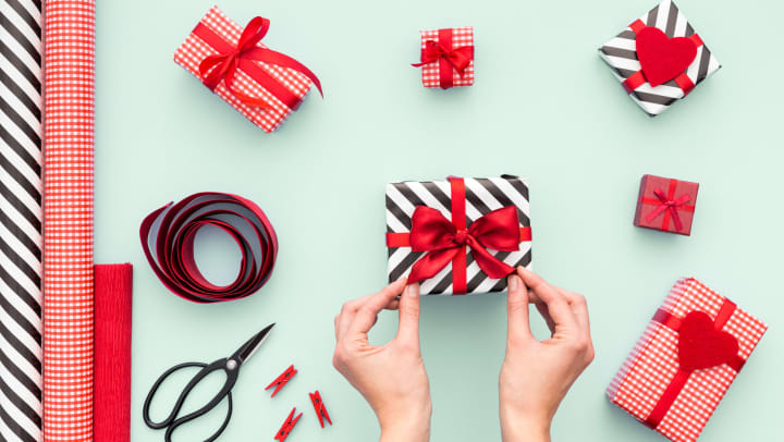 Hands holding the ends of a red bow on a wrapped package with other wrapped gifts surrounding it. 