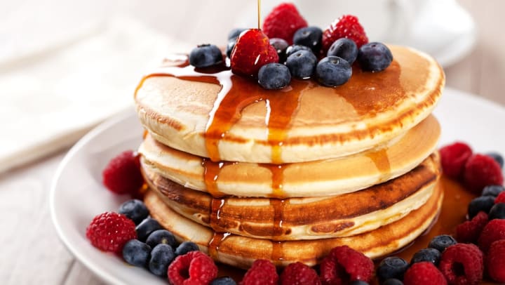 A stack of four fresh pancakes on a plate, topped with blueberries and raspberries, as well as syrup and a pad of butter