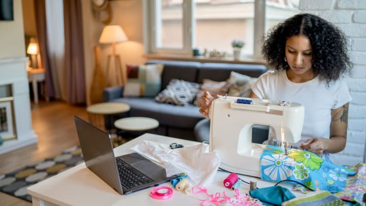 A woman sits at her kitchen table while using a sewing machine to stitch fabric with a bright blue floral design | craft stores in Jacksonville