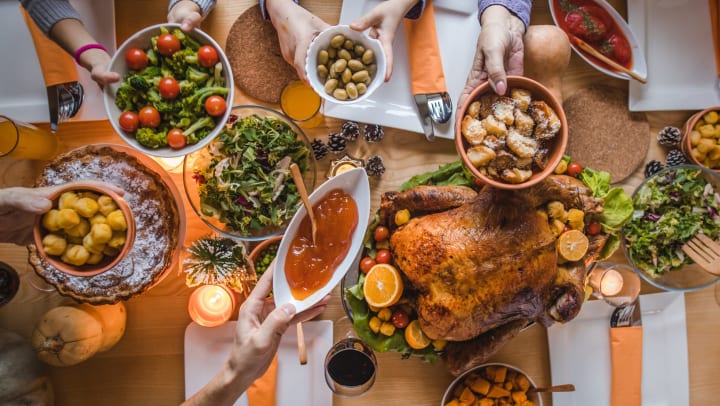 Hands holding up various side dishes above a dining table full of food, including a giant turkey. 