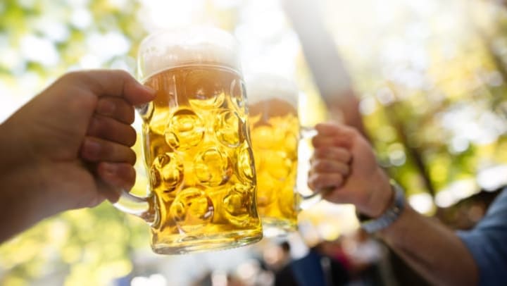 View of two hands holding glasses of beer and clinking them together in a bright outdoor setting with green trees and a group of people in the background