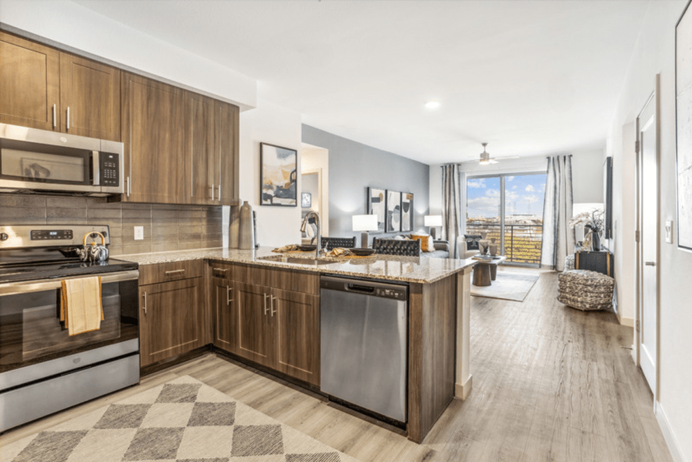 Kitchen with stainless steel appliances, quartz countertops, and living room with wood-style flooring and balcony atThe Margaret at Riverfront in Dallas, Texas