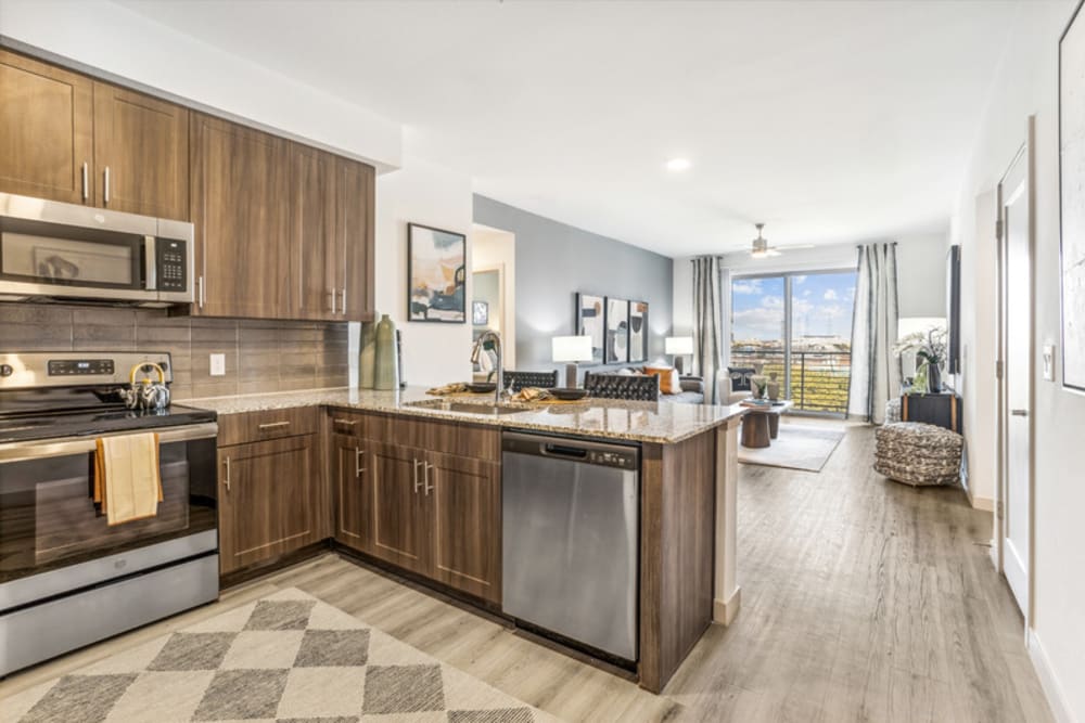 Kitchen with stainless steel appliances, quartz countertops, and living room with wood-style flooring and balcony atThe Margaret at Riverfront in Dallas, Texas
