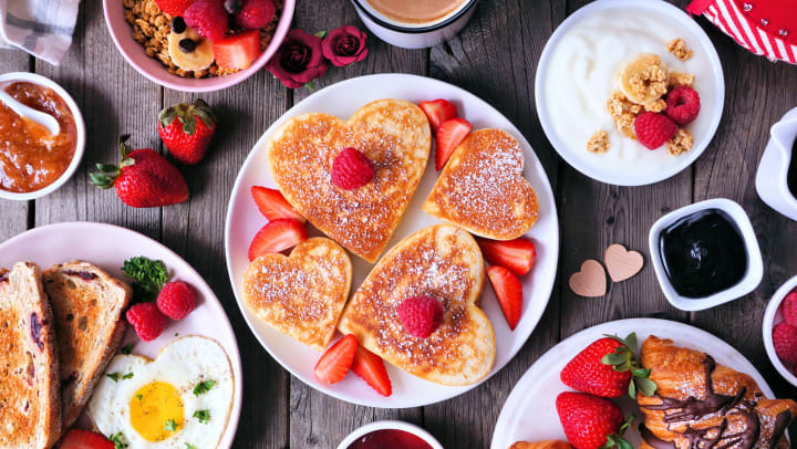  Various breakfast foods on a table with heart-shaped pancakes in the center with raspberries and strawberries on top. 
