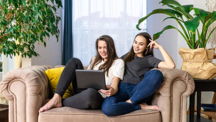 Two young women sit on a sofa looking at a laptop, surrounded by plants and a macrame wall hanging. 