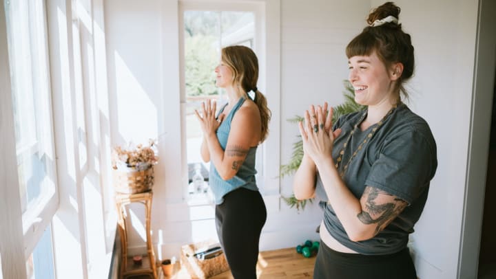 Two smiling young women standing on yoga mats while practicing yoga in a bright sunlit interior