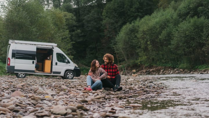  A forest and open van in the background. Rocky beach next to a stream. A man and a woman are sitting together on the rocky beach. 