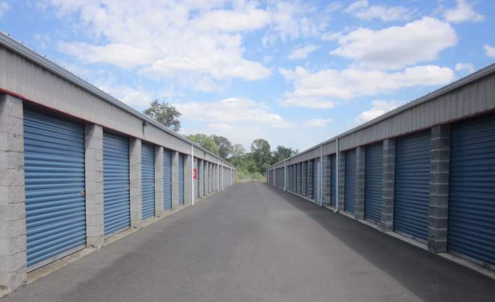 a view of a storage facility facing down a wide and easy to access aisle with blue storage doors in a concrete building with a blue sky full of puffy white clouds at the top