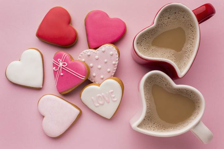 an above photo of heart shaped cookies with various red, pink, white, and polka dot frosting, one cookie spells the word LOVE, beside two heart shaped coffee mugs