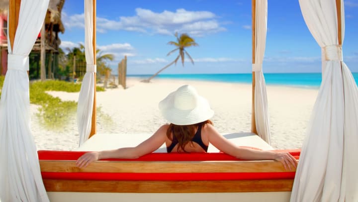 Woman reclining on a padded bench in a cabana on the beach, with her back to the camera.