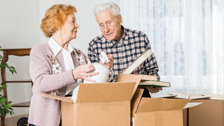 An older man and woman are lifting boxes into a room, suggesting they are unpacking or settling into a new environment