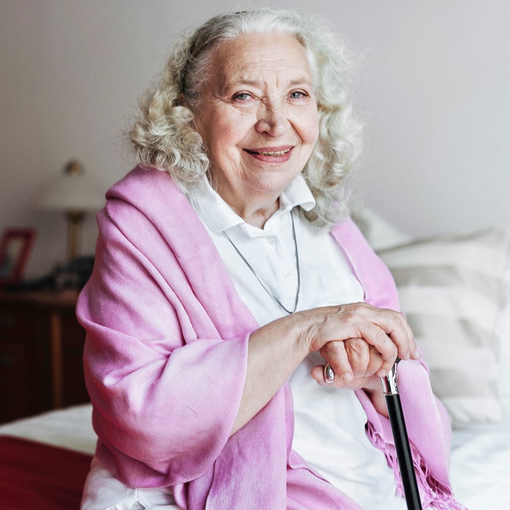 A resident with a cane sitting on the edge of her bed at Chateau Gardens Memory Care in Springfield, Oregon