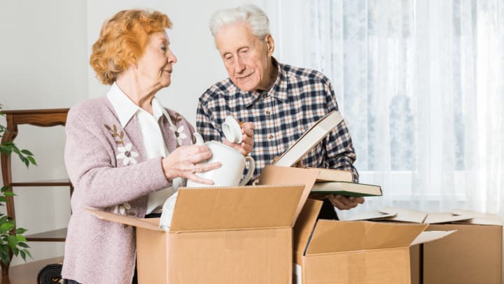 An older man and woman are lifting boxes into a room, suggesting they are unpacking or settling into a new environment
