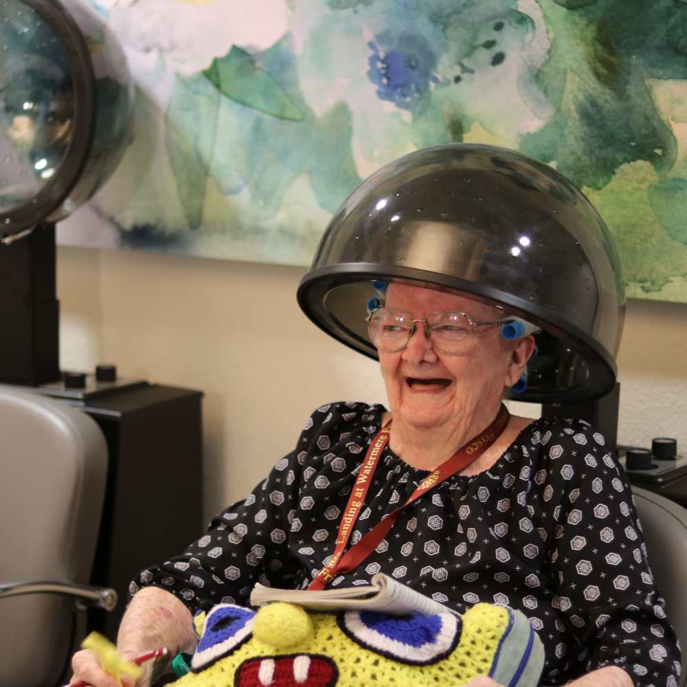 Senior woman laughing while under a hair dryer during a salon visit at Watermere Frisco memory care community.