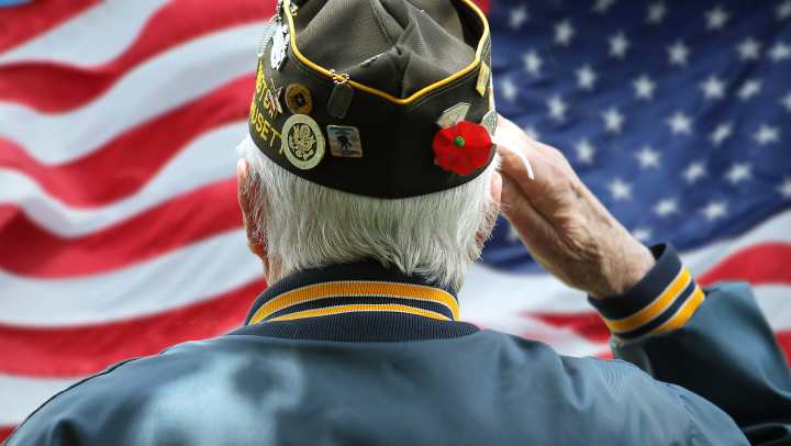 An elderly U.S. military veteran salutes the American flag, wearing a decorated cap and jacket with service insignia. A heartfelt tribute to veterans and their service.