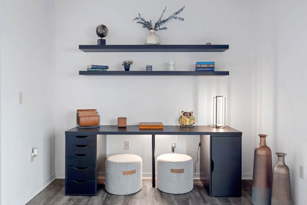 Modern workspace nook with a black desk, storage drawers, decorative shelves with books and plants, two cushioned stools at Carlton Arms Of Magnolia Valley in New Port Richey, Florida