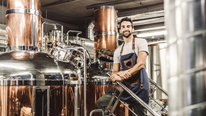 Man standing in front of polished copper and chrome distilling equipment, facing the camera and smiling. 