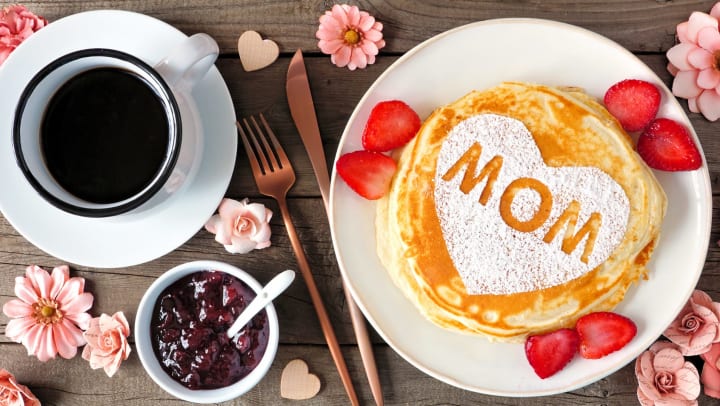  Pancake with powdered sugar spelling mom on it, surrounded by jam and coffee on a table decorated with pink flowers. 