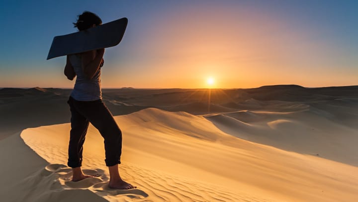 Person standing atop a sand dune, holding a sandboard up at their shoulders
