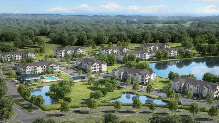 An aerial architectural rendering of Alera at Hunters Glen Apartments in Lake Wales, FL. The image shows several multi-story apartment buildings with gray and white siding clustered near two small ponds and a larger lake, surrounded by abundant green grass and dense forest. A swimming pool and clubhouse area are visible in the center-left. The buildings are laid out with parking lots and connecting roads on a sunny day.
