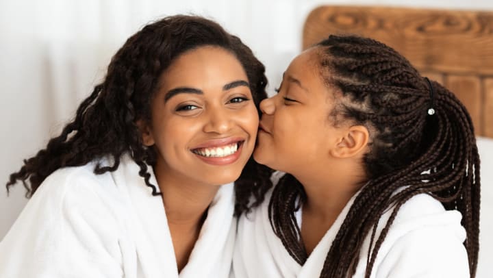  Young girl kissing an adult woman on the cheek who is smiling, both clothed in white robes. 